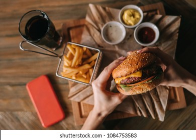 Woman holds burger with hands and fries on the background in cafe. Fresh burger cooked at barbecue in craft paper. American food. Big hamburger with meat and vegetables closeup unfocused at background
