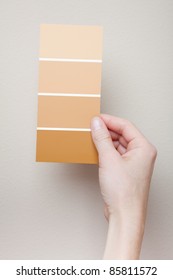 A Woman Holds A Card With Four Paint Swatches Near A Wall For Comparison.