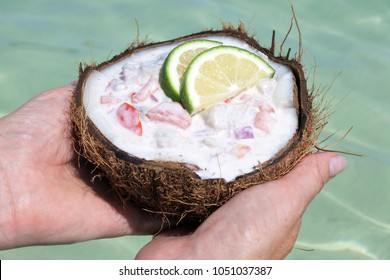 Woman Holds Ceviche Dish In A Coconut Over Lagoon Water In Rarotonga, Cook Islands. Food Background And Texture. Copy Space