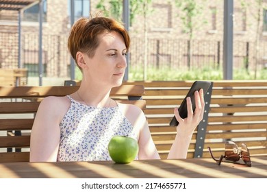 Woman Holds An Ebook Device And A Reusable Coffee Cup In Her Hands. A Woman Reads A Book Near The Office During A Work Break. Lunch Break On A Summer Day.