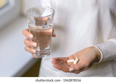 A woman holds a glass of water and two pills in her hands. Young woman treats illness with pills. Headache or stomach problems and treatment with medication. Antibiotic treatment concept - Shutterstock ID 1963878556