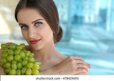 Woman holds grapes in the chest and hugs it with both hands. Beauty portrait with bare shoulders and her hair gathered against the glass background