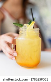 Woman Holds In Hand Delicious Yellow Lemonade With Ice And Fresh Mint On A White Table. Summer Healthy Fruit Drink. Close Up.