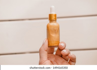 Woman Holds In Hand One Glass Cosmetic Bottle With Dropper Pipette. Luminous, Luxurious Gold Liquid Highlighter. Beauty Blogger Skincare Essentials. Minimalism Branding Package. Overhead. Copyspace