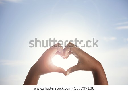 Woman holds hands up to sky in the shape of a heart around the sun. 