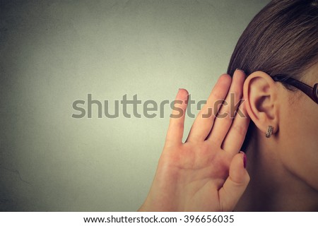 Woman holds her hand near ear and listens carefully isolated on gray wall background 