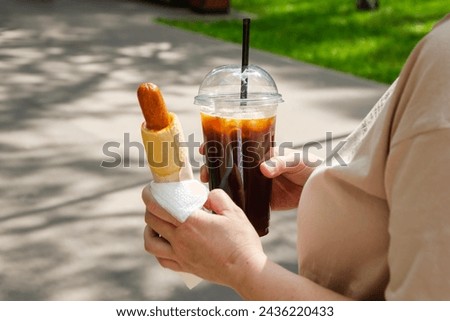 woman holds a hot dog and black ice coffee in her hands while walking in the park. Street food