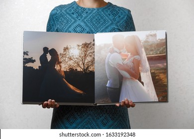 A Woman Holds Open Family Photobook. The Person Looks At The Wedding Photo Book. Sample  Photo Album. Wedding Photoalbum With Fabric Cover. Female Hands Holding Square Open Wedding Photo Album.