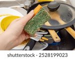 Woman holds orange sponge for washing dishes. The kitchen sink is full of dirty dishes. An old dirty kitchen sponge