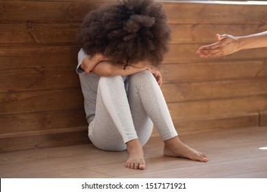 Woman holds out her hand to crying abandoned african child, little girl sit on floor closed with hands weeping, new mom for orphan kid, racial discrimination, mental problem and giving support concept