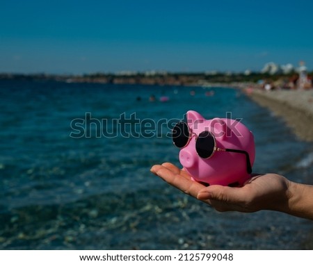 A woman holds a piggy bank in sunglasses on a pebble beach near the sea. Budget vacation.