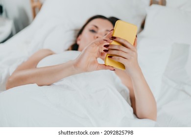 Woman holds smartphone in hands while lying in comfortable bed. Lady with mobile in light cozy bedroom. Addiction to technology and social media, morning rituals concept. High quality photo