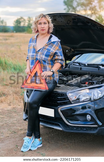 A woman holds a\
triangular emergency stop sign in her hand while standing at the\
open hood of a broken car