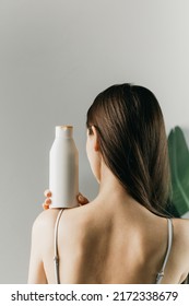 Una mujer sujeta una botella blanca de cosméticos en su mano, de pie con la espalda a la cámara. Muéstrate champú, gel.