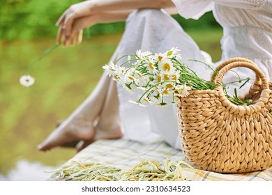 a woman holds a wicker bag with daisies with her hand while sitting on the pier