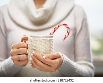 Woman holds a winter cup close up. Woman hands with elegant french manicure nails design holding a cozy knitted mug with cocoa, tea or coffee and a candy cane. Winter and Christmas time concept. 