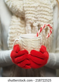 Woman holds a winter cup close up. Woman hands in woolen red gloves holding a cozy mug with hot cocoa, tea or coffee and a candy cane. Winter and Christmas time concept.  