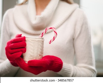 Woman holds a winter cup close up. Woman hands in woolen red gloves holding a cozy mug with hot cocoa, tea or coffee and a candy cane. Winter and Christmas time concept.
