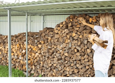 A woman in home clothes gathers firewood for heating from a woodpile. Heating the house with wood. The girl stands with her back and picks up firewood - Powered by Shutterstock