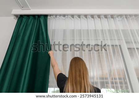 A woman at home covers the thick green curtains hanging on the curtain rod