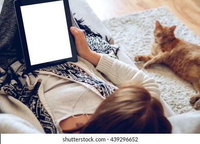 Woman in home cozy clothes lying on a sofa using tablet with headphones, looking at a lazy red cat sprawled on the carpet beside. Online education concept. e-learning. back view. Pet shop concept