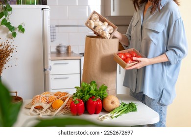 Woman at home getting groceries out of a shopping bag with grocery ordered from internet. Fresh organic vegetables, greens and fruits. Kitchen interior.  Food delivery concept - Powered by Shutterstock