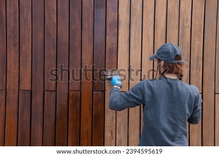 Woman home owner applying protective varnish or paint on wooden house wall cladding