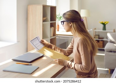 Woman at home pulls out of envelope document she received by mail or delivery service. Side view of woman sitting at table reading letter with paper invoice or receiving bank receipt for tax refund. - Shutterstock ID 2075286412
