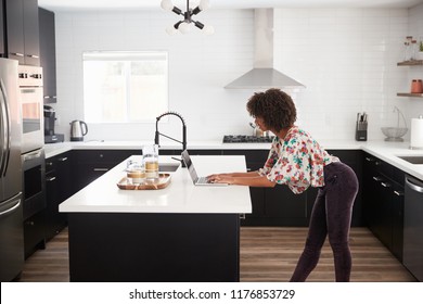 Woman At Home Standing At Kitchen Island Whilst Using Laptop