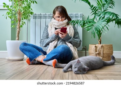Woman at home using smartphone, warming with cat near heating radiator