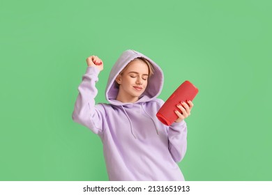 Woman in hoodie with wireless portable speaker enjoying music and dancing on green background