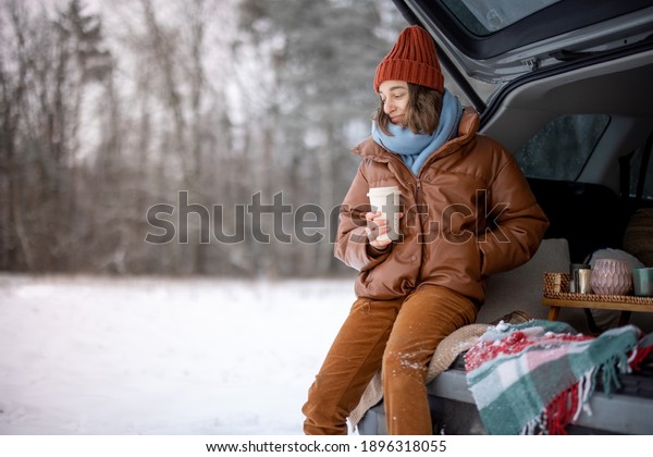 Woman\
with hot tea sitting in car trunk and look at the forest, traveling\
by car during winter holidays. High quality\
photo