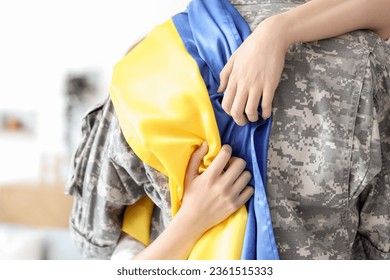 Woman hugging male soldier with flag of Ukraine at home, closeup