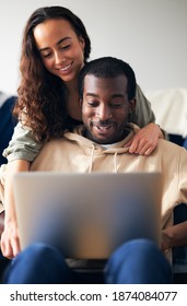 Woman Hugging Man From Behind As Young Mixed Ethnicity Couple Relax On Sofa At Home Browsing Internet On Laptop