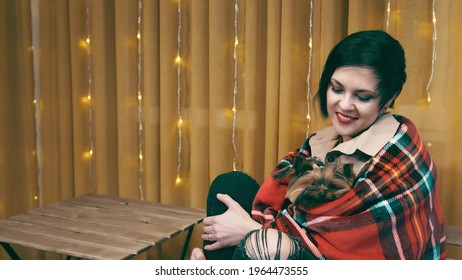 The Woman Hugs And Warms Her Yorkshire Terrier Dog. A Small Dog Looks Out From Under A Warm Blanket. Family Comfort And Warmth With Pets.