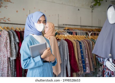 A Woman In A Hveil Wearing A Mask In Blue Holding A Pad Is Standing Next To A Mannequin In A Muslim Clothing Shop