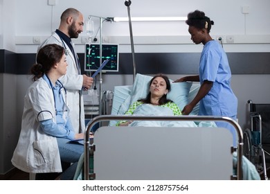 Woman With Illness Connected To Iv Drip Line And Monitor Showing Vitals Recieving Medical Care From Nurse. Team Of Doctors Do Clinical Consult On Patient With Nasal Cannula Recovering From Surgery.