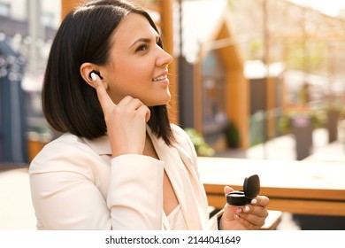 Woman inserts earbuds into her ear and turns them on to listen to music or for the convenience of communication. Lady on the street in the patio of the city cafe. Wireless modern headphones in a box