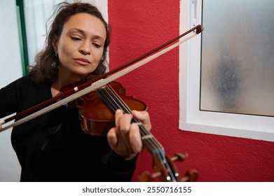 Woman intently playing the violin, creating serene music indoors next to a window. The red wall adds a vibrant backdrop to her focused expression, highlighting a moment of passion and artistry. - Powered by Shutterstock