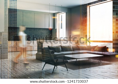 Woman in interior of modern living room with gray walls, wooden floor, gray sofa and armchair standing near coffee table and kitchen in the background. Toned image double exposure blur
