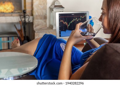 woman investing in a stock exchange from the living room of her house, with the fireplace lit and drinking a glass of wine - selective focus
