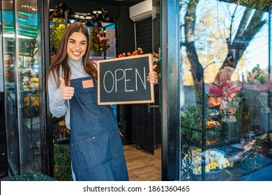 Woman Inviting To Her Flower Shop. Beautiful Smiling Young Florist In Apron Holding Open Sign. Smiling Woman Holding Open Sign, Written On Blackboard With Chalk, Outside Of Flower Shop.