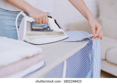 Woman ironing blue shirt with steam iron next to stack of washed and folded cotton shirts on ironing board after laundry. Housekeeping concept