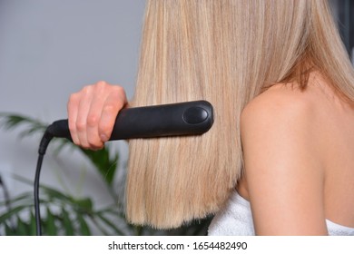 Woman Ironing Long Blonde Hair With A Flat Iron At Home. Cares About Healthy And Clean Hair. Girl Wears A White Towel. Female With Bare Shoulders. Beauty Salon Concept. Close Up, Selective Focus