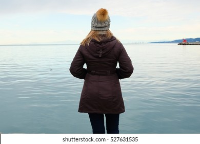 Woman in jacket and hat looking at the sea during winter walks on the beach, view from the back