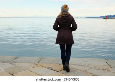 Woman in jacket and hat looking at the sea during winter walks on the beach, view from the back