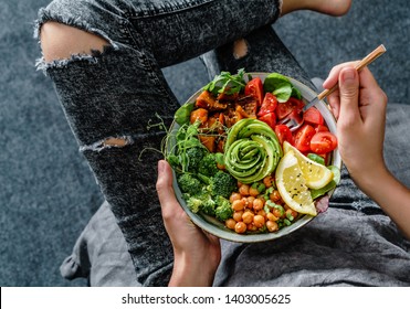 Frauen in Jeans, die Buddha Schüssel mit Salat, gebackene Süßkartoffeln, Hühnererbsen, Brokkoli, Grün, Avocado, Sprossen in den Händen halten. Gesunde veganische Lebensmittel, sauberes Essen, Diät, Draufsicht
