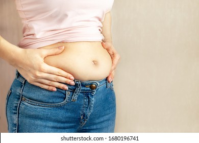 Woman in jeans and a light shirt is standing sideways and holding hands squeezes excess belly fat on light background, copy space. The concept of overweight, weight loss, diet, obesity, junk food.