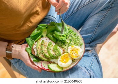 A Woman In Jeans Sits And Holds A Plate With Fresh Food Avocado, Cucumber, Spinach, Radish, Egg, Microgreens, Seeds. Proper Healthy Eating, Keto Diet. Green Vegan Breakfast. Close-up Selective Focus