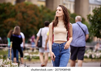 Woman In Jeans And Top Walks In Crowded City Park On Summer Evening.